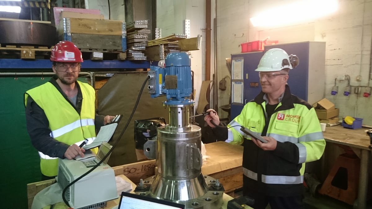 Two men in hard hats and high visibility clothing at a workshop bench, taking measurements on the Lynx 250: a stainless steel housing with a blue electric drive motor mounted on top.