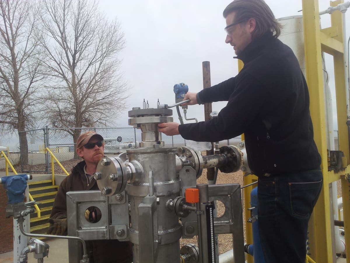 Two men at a stainless steel separator built into an outdoor test loop, with bolted flanges and instrument lines. One of them reaches over the top of the unit.