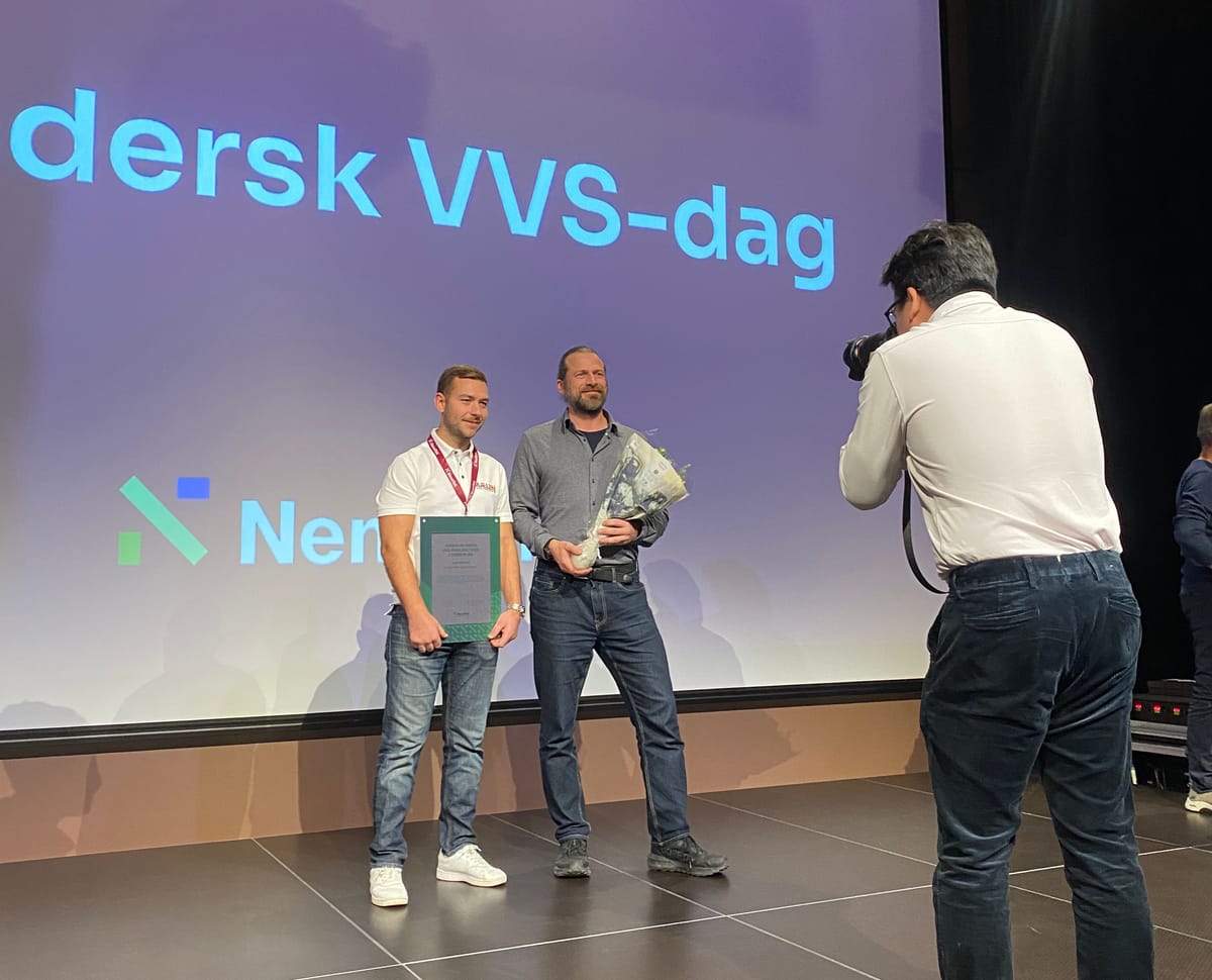 Two men on a stage holding a framed diploma and a bouquet, in front of a screen showing the NemiTek logo, while a photographer takes their picture.
